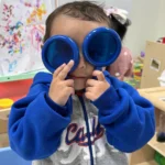 A child exploring with toy binoculars in the STEM learning area of the GMCC classroom