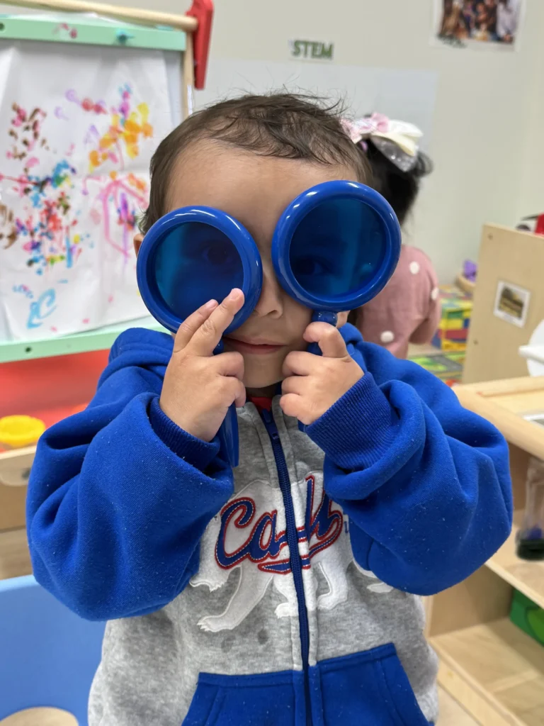 A child exploring with toy binoculars in the STEM learning area of the GMCC classroom