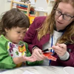 A teacher guiding a toddler through a hands-on painting activity at GMCC early learning center