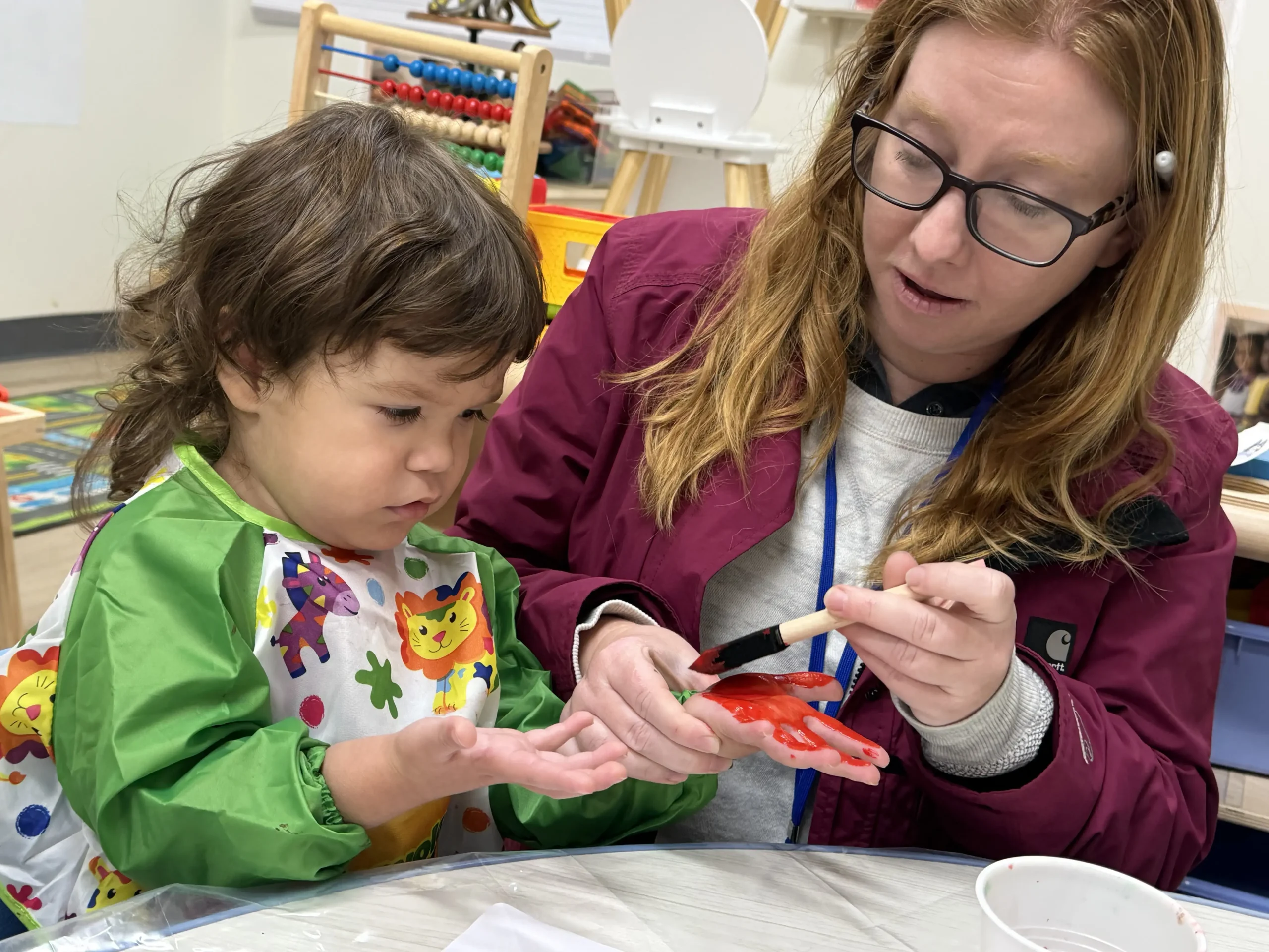 A teacher guiding a toddler through a hands-on painting activity at GMCC early learning center