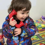 A toddler enjoying pretend play with a toy telephone in the GMCC classroom