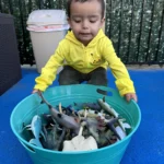 A toddler exploring a bucket of sea animal toy figures during sensory play at GMCC